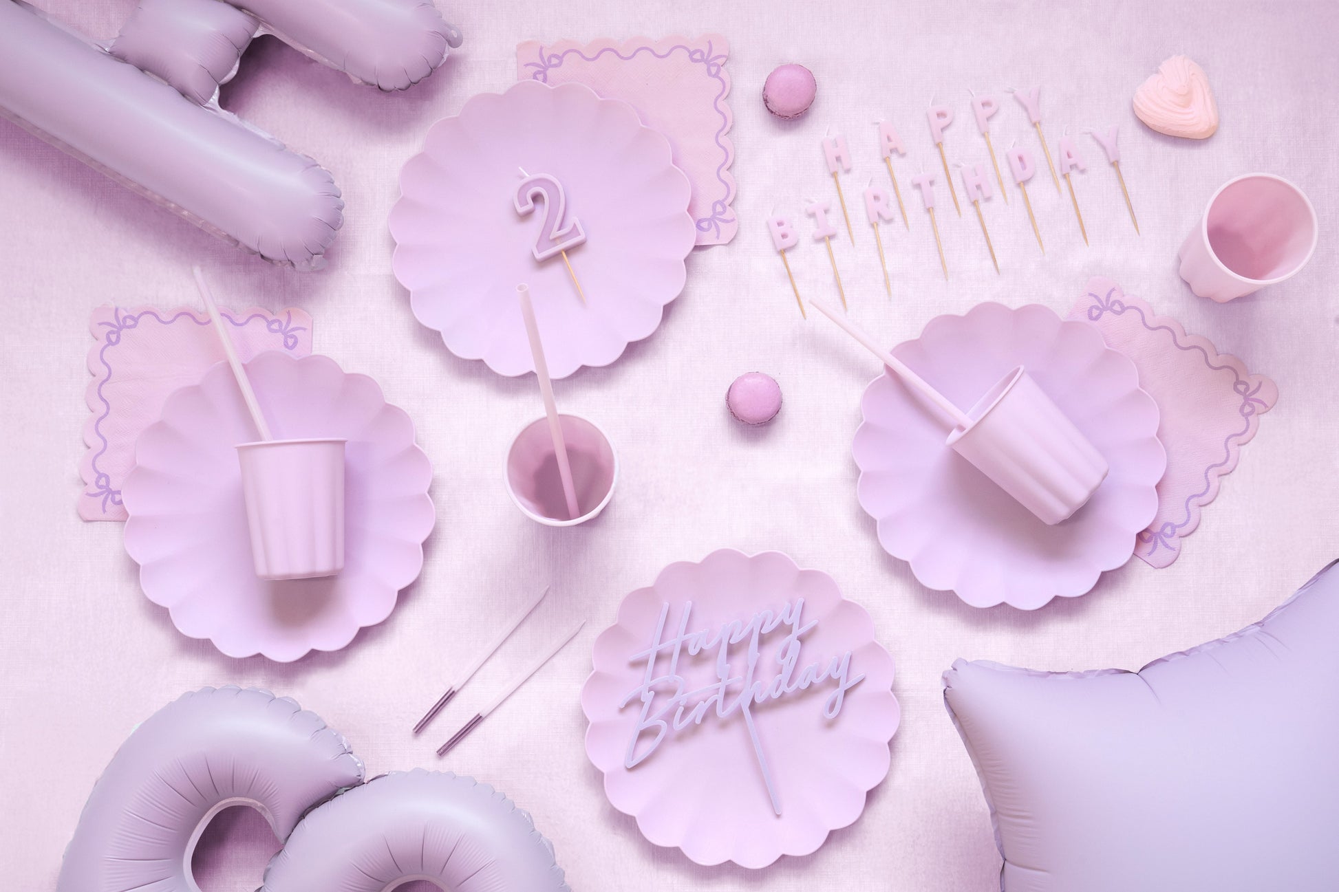 Pink and purple birthday party supplies including plates, cups, and balloons on a pink background.