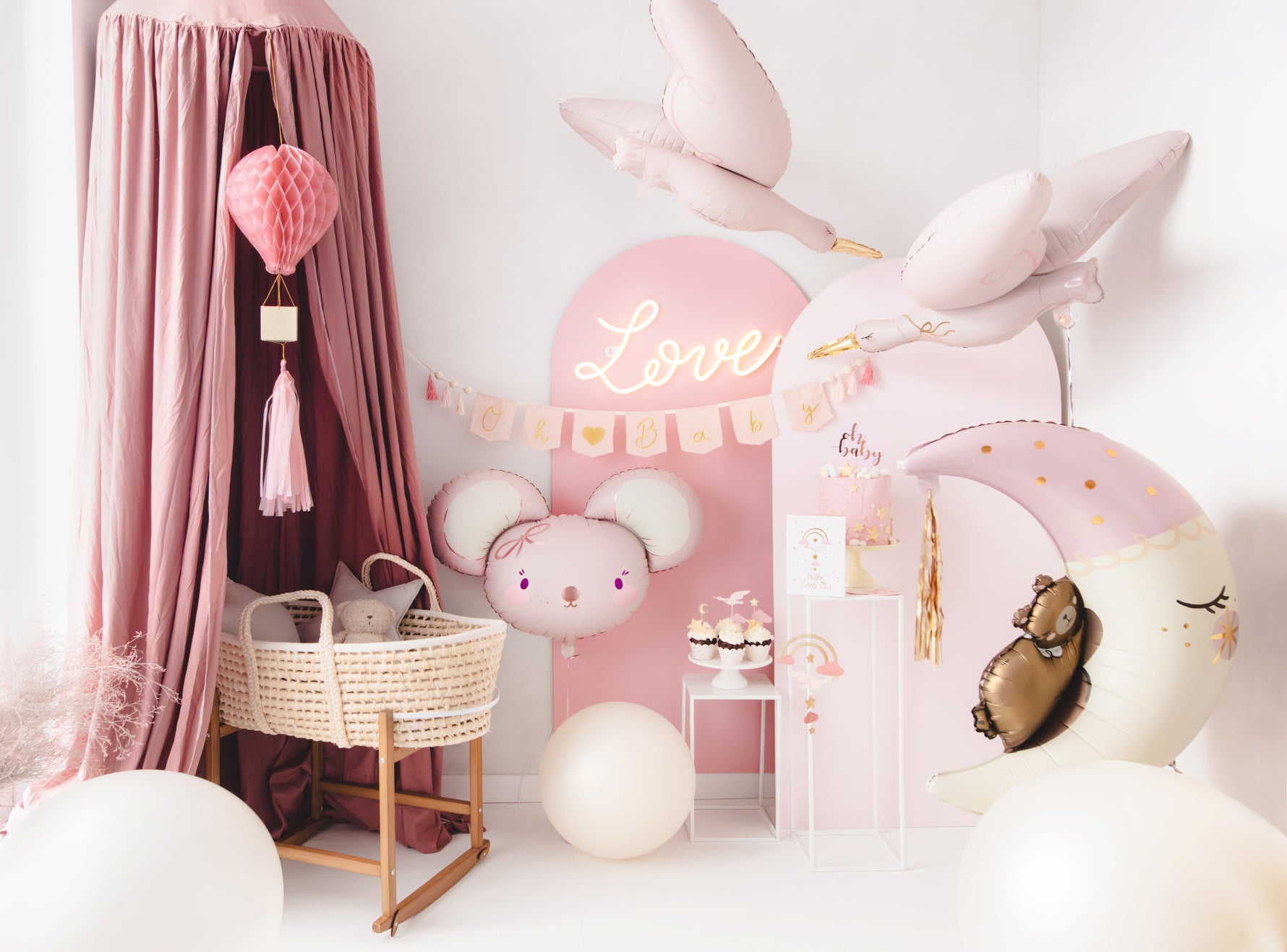 Baby girl welcome party setup in soft pink tones with stork and moon foil balloons, Oh Baby banner, mouse balloon, cake display, and bassinet under canopy – perfect for a newborn celebration.