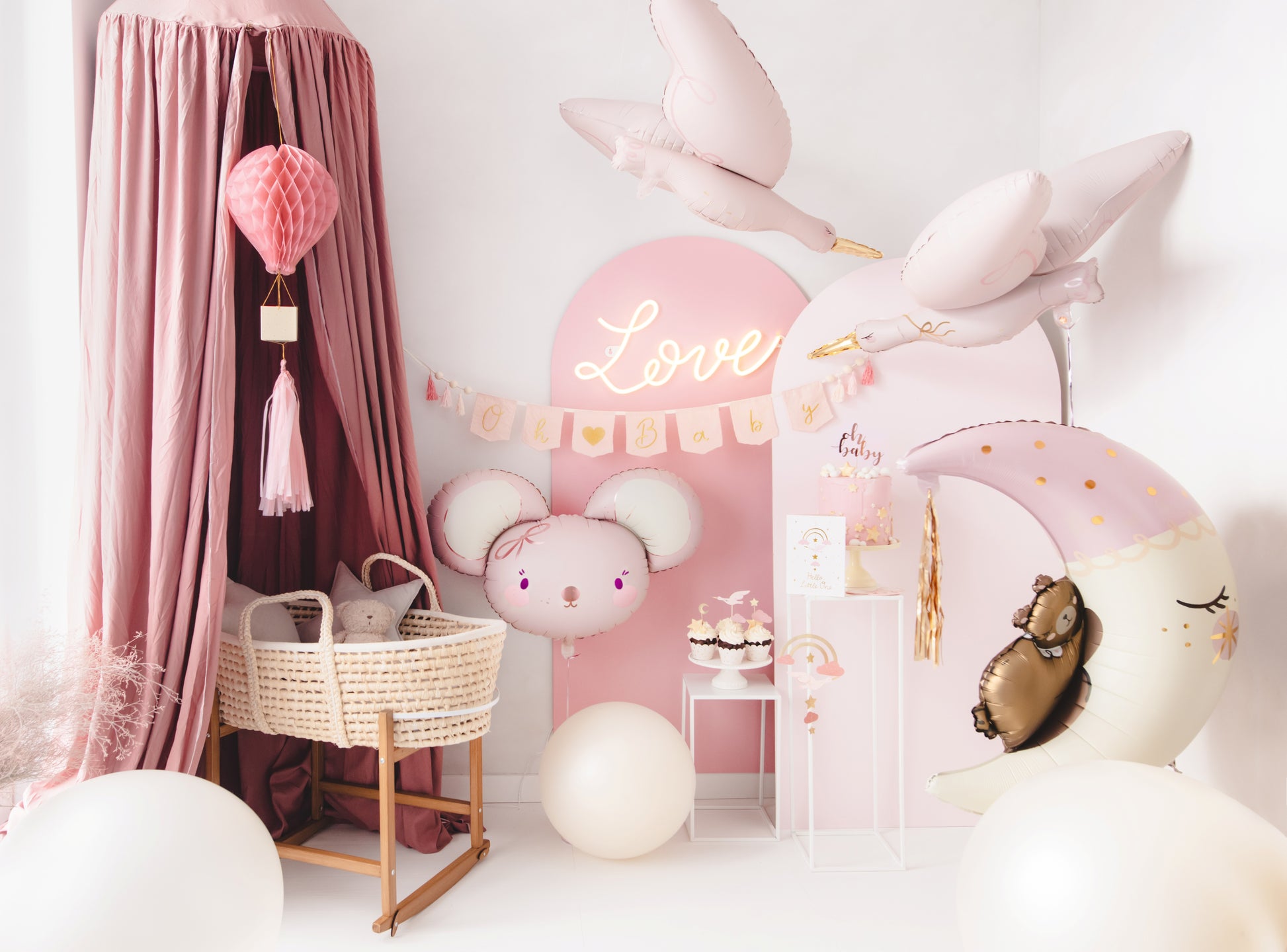 Baby girl welcome party setup in soft pink tones with stork and moon foil balloons, Oh Baby banner, mouse balloon, cake display, and bassinet under canopy – perfect for a newborn celebration.