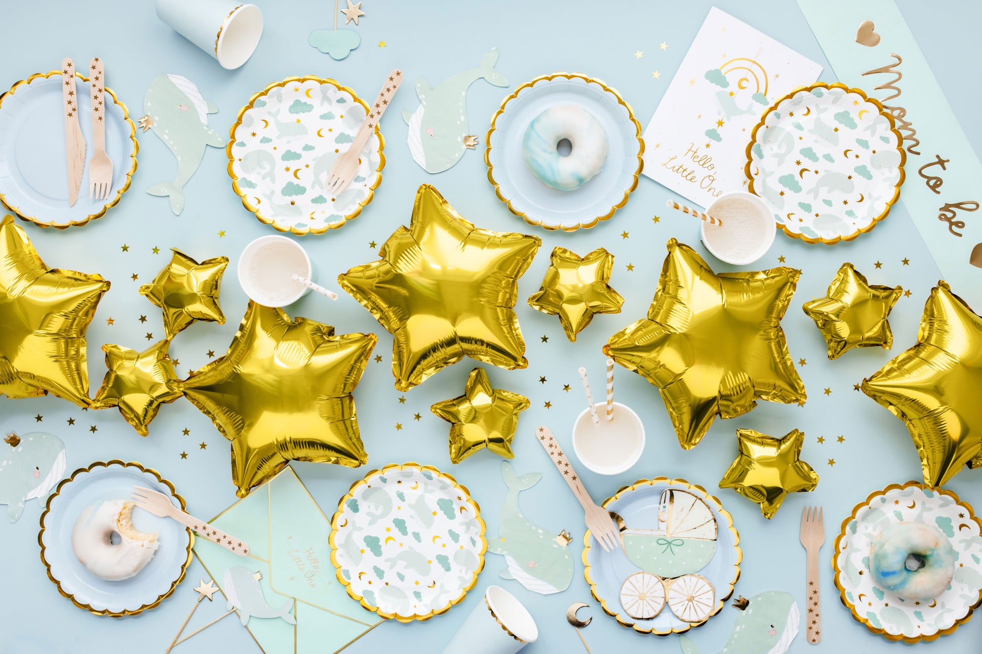 Flat lay of pastel blue baby shower table setup with golden star balloons, cloud and whale themed paper plates, cups, and wooden cutlery – perfect for a 'Welcome to the World' baby boy celebration.