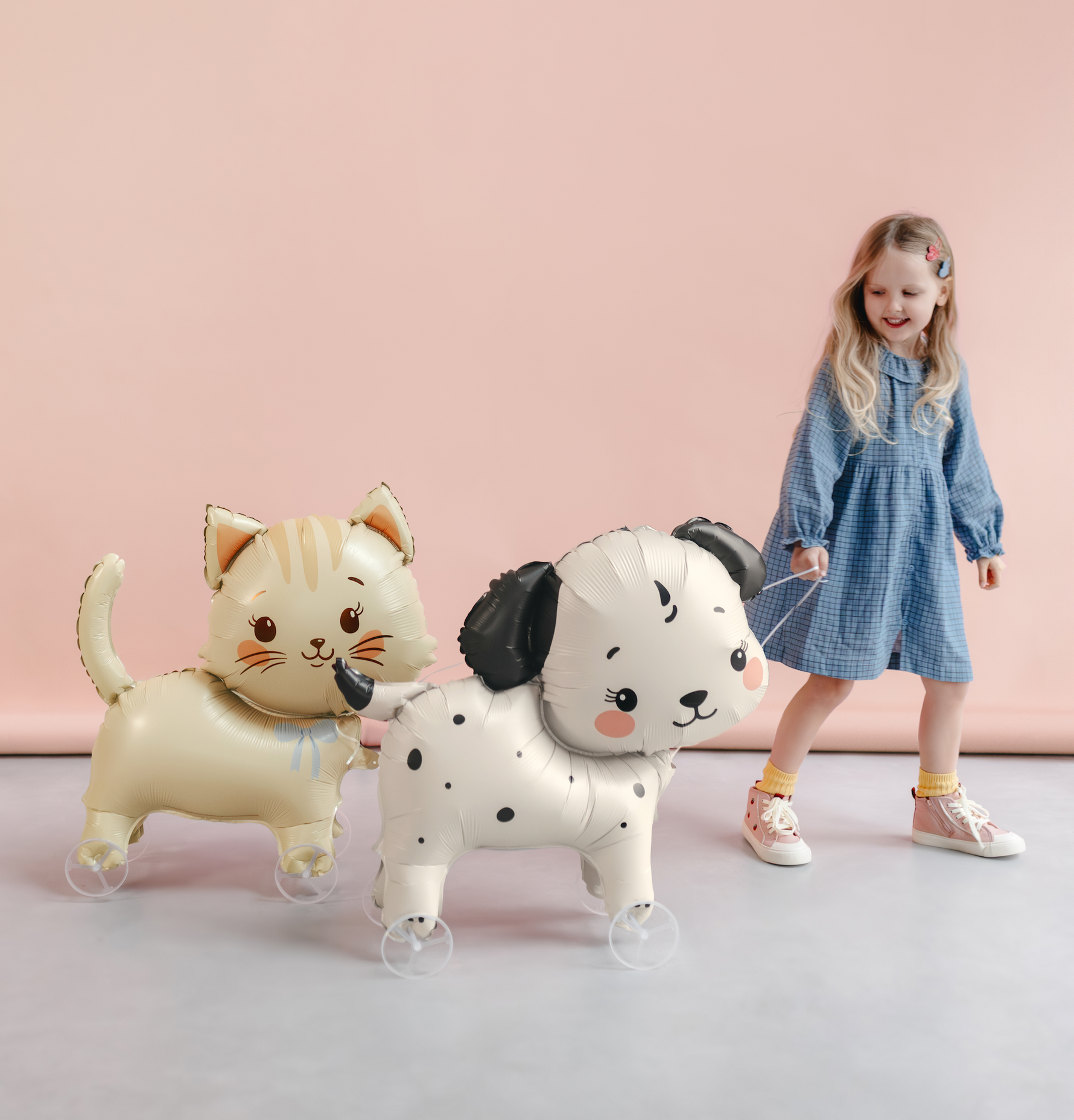 A young girl in a blue dress stands on a light gray floor against a soft pink wall, holding a string attached to two large inflatable balloon animals: a cream-colored cat and a white dog with black spots.
