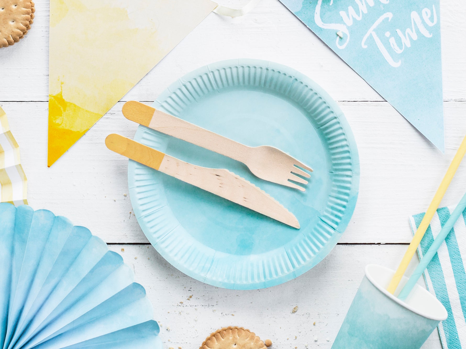 Light blue paper plates with woden cutlery on a white wooden surface with blue party decorations.