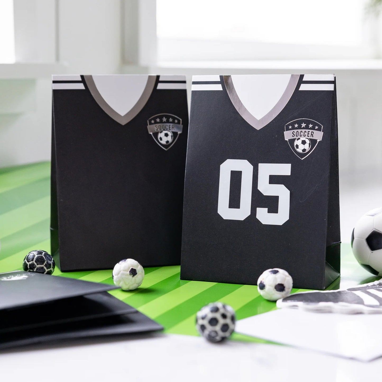 Two black soccer jersey-themed treat bags on a green surface with soccer balls around.