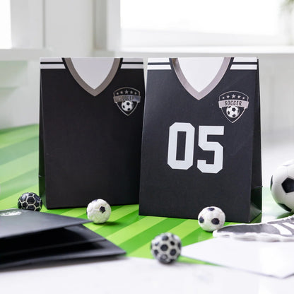 Two black soccer jersey-themed treat bags on a green surface with soccer balls around.