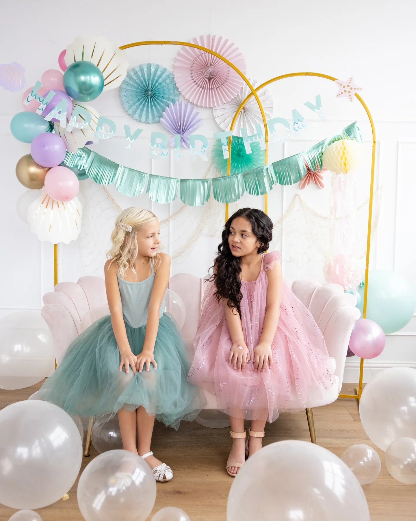 Two young girls in tutu dresses sitting under a decorative arch with balloons and streamers.