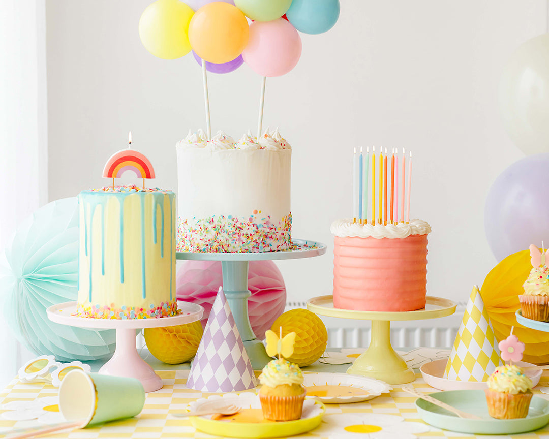 Colorful birthday cake display with pastel balloons and party hats on a checkered tablecloth.