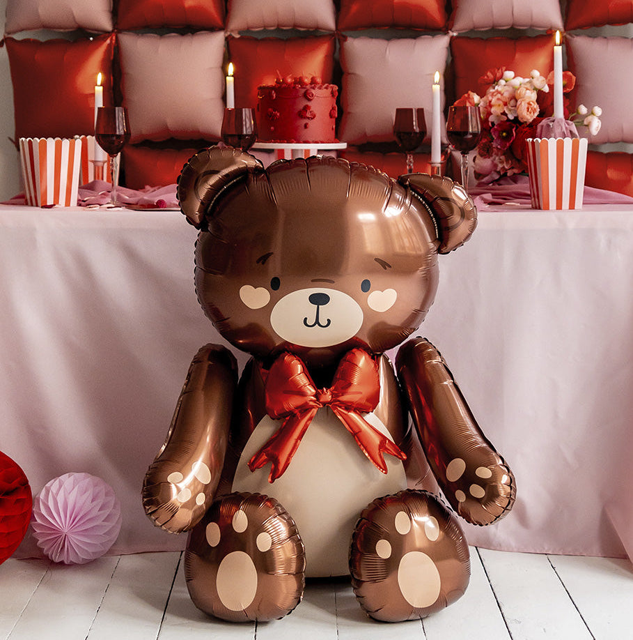 Brown teddy bear-shaped balloon with a red bow tie on a festive table.