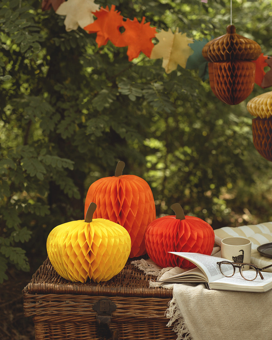 Three honeycomb paper pumpkin decorations in yellow, orange, and red are arranged on a wicker trunk outdoors, with a tablecloth, open book, glasses, and hanging acorn ornaments in the background.