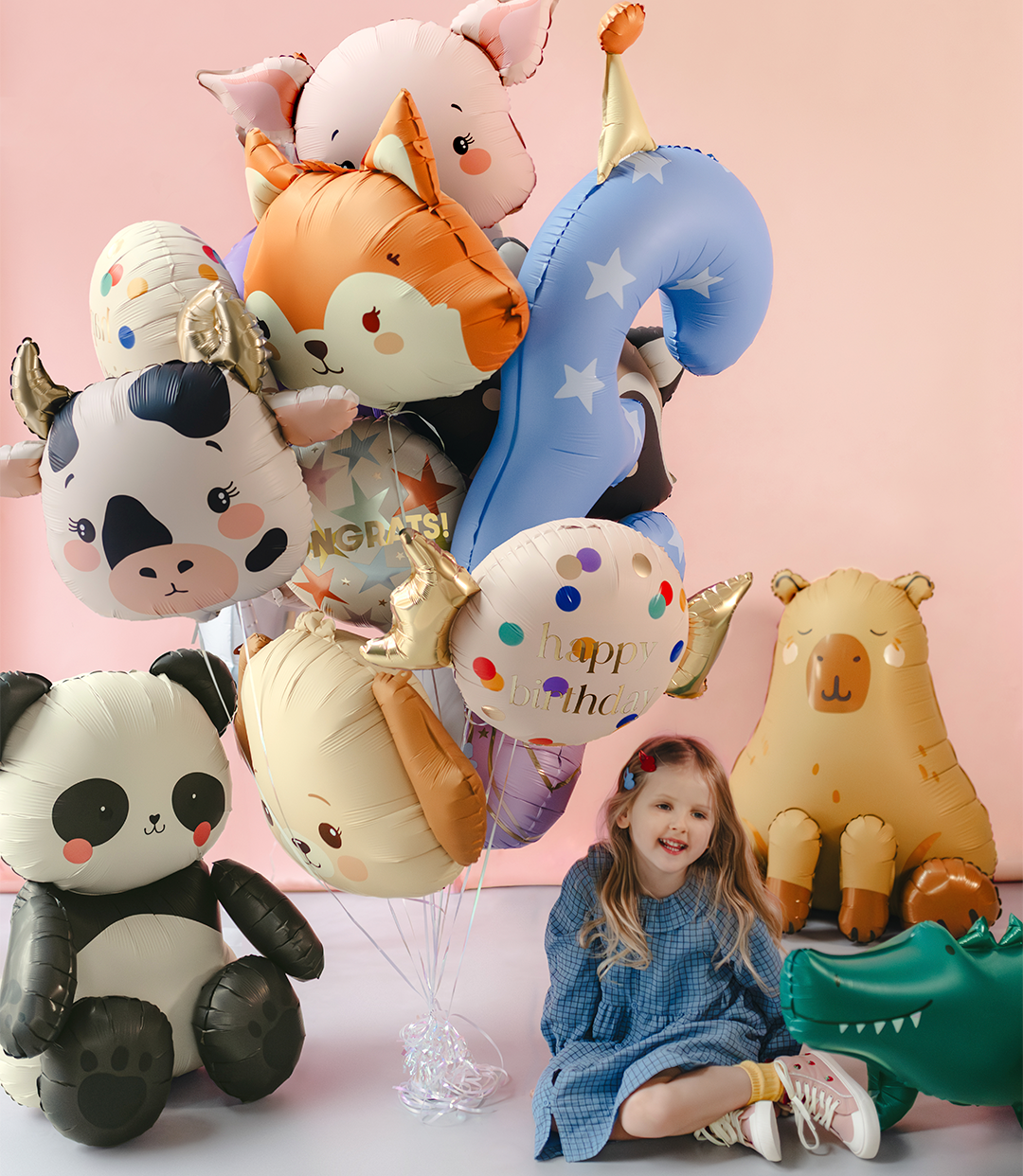 Child sitting among animal-shaped balloons against a pink background