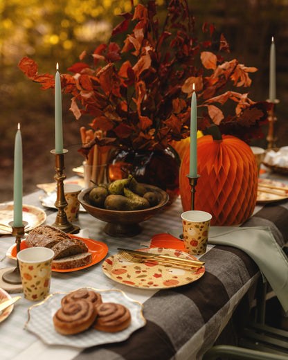 An autumn-themed table setting with a large orange honeycomb pumpkin, a dark glass vase filled with red and orange fall leaves, a bowl of pears, cinnamon rolls, patterned paper plates and cups, and lit teal taper candles on a checkered tablecloth.