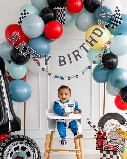 Child in a high chair under a 'Happy Birthday' balloon arch with colorful balloons and decorations.