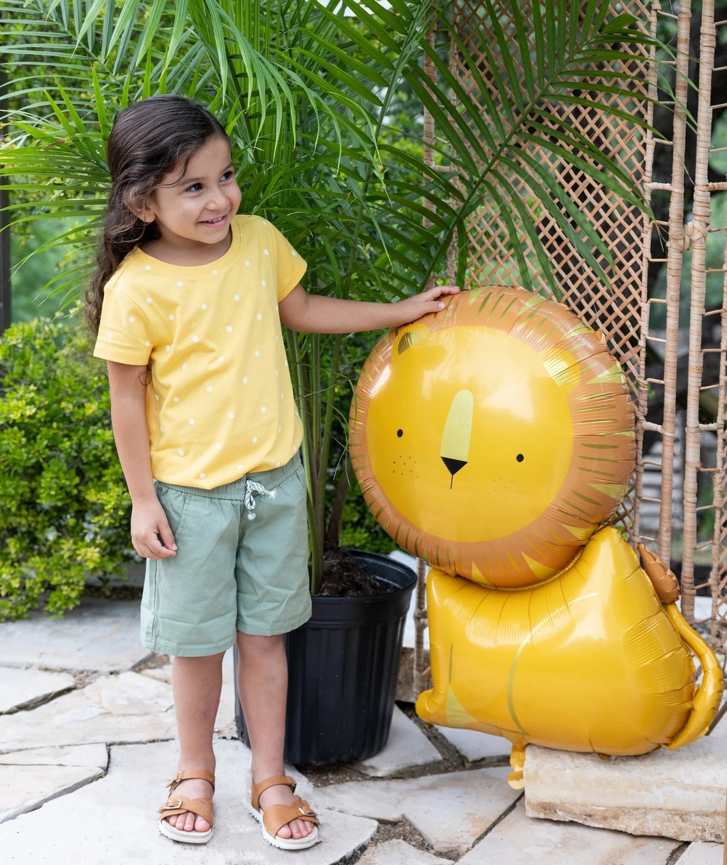 Child in yellow shirt and green shorts standing next to a lion-shaped balloon outdoors.