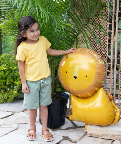 Child in yellow shirt and green shorts standing next to a lion-shaped balloon outdoors.